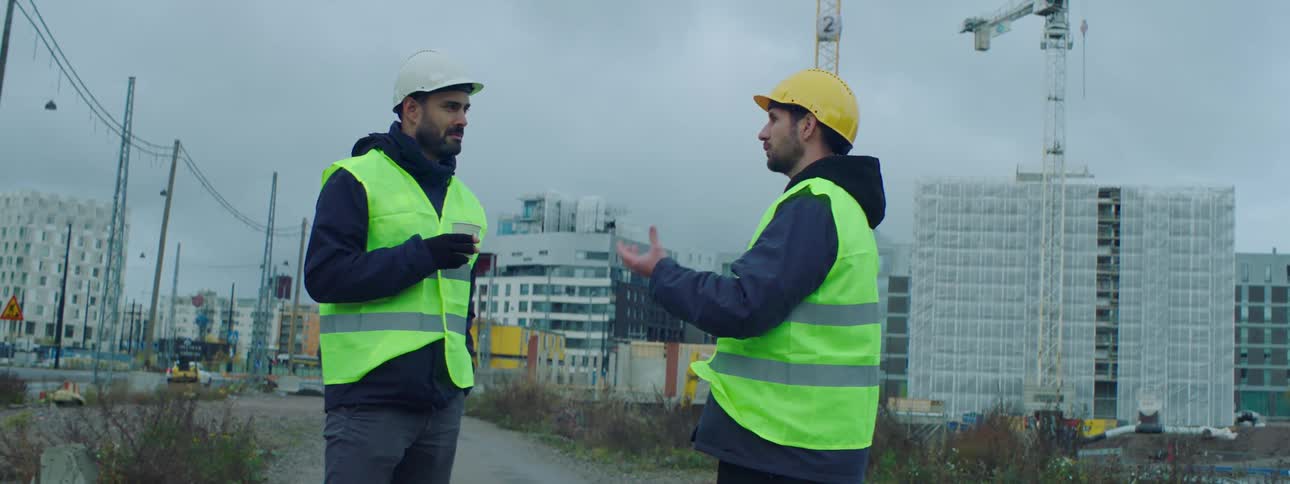 Two construction workers are standing outside and one is holding a cup and the other is talking