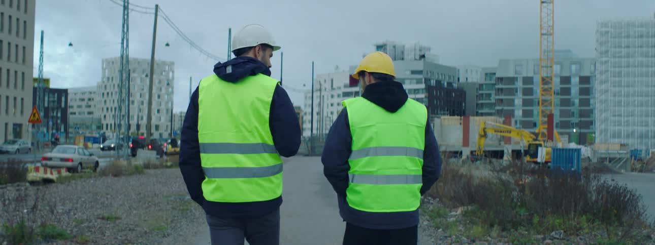 Two white men walking together and talking at a construction site wearing safety helmet and vests