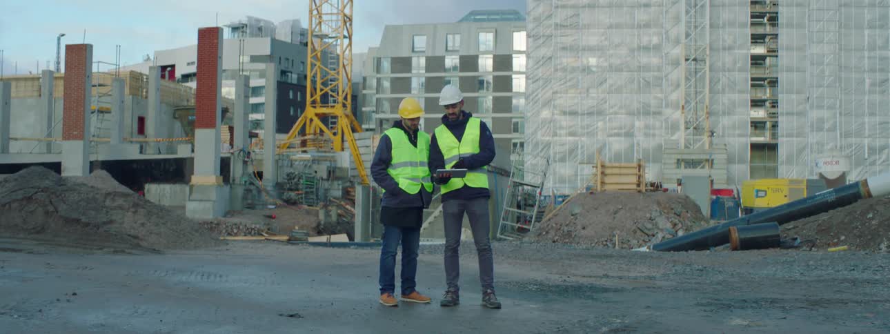 Two male construction workers wearing safety helmets and green vests standing in the middle of a construction site using a tablet