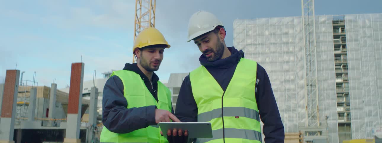 Construction worker holds a tablet as he and a coworker look at it and talk