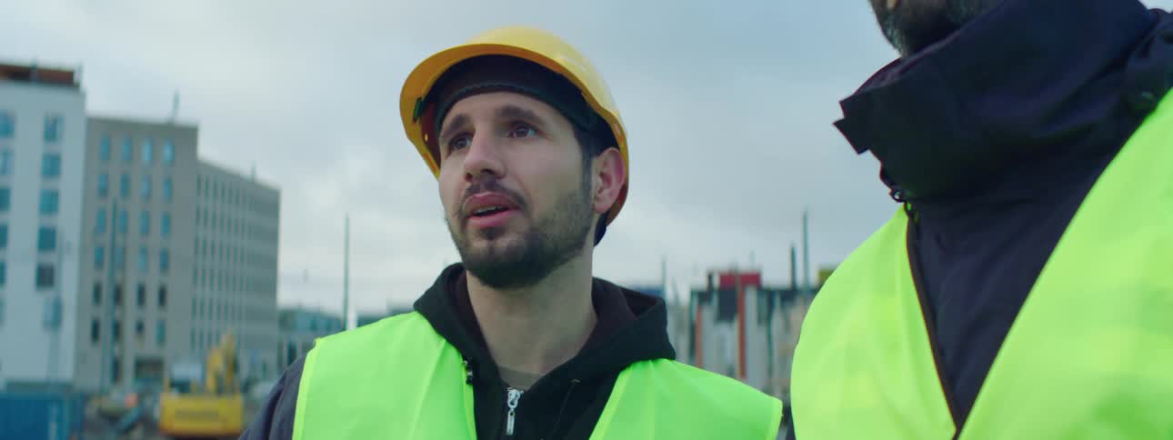 Two construction workers wearing bright yellow vests and hardhats are standing outside talking