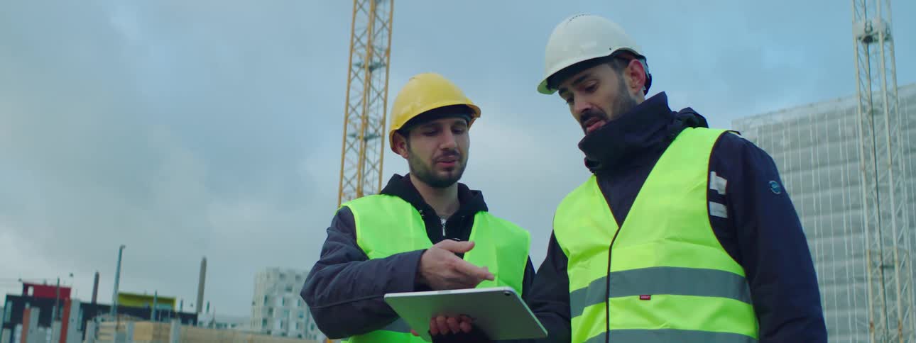 Two construction workers talk at a job site while holding a tablet