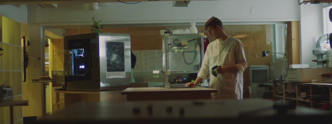A man wearing a white lab coat is looking at a machine and then walks over to a laptop computer sitting on a table