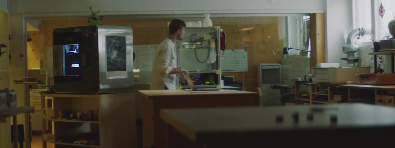 A man wearing a white lab coat is operating a 3d printer and then he sits at a table with a laptop computer