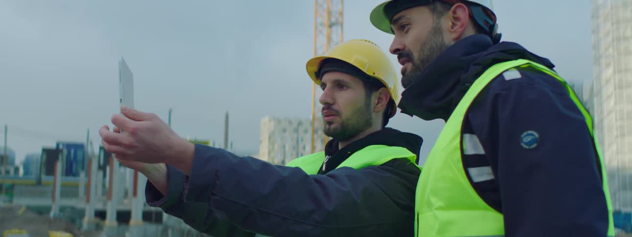 A construction worker holding up a tablet in outstretched hands at a site talking with another man