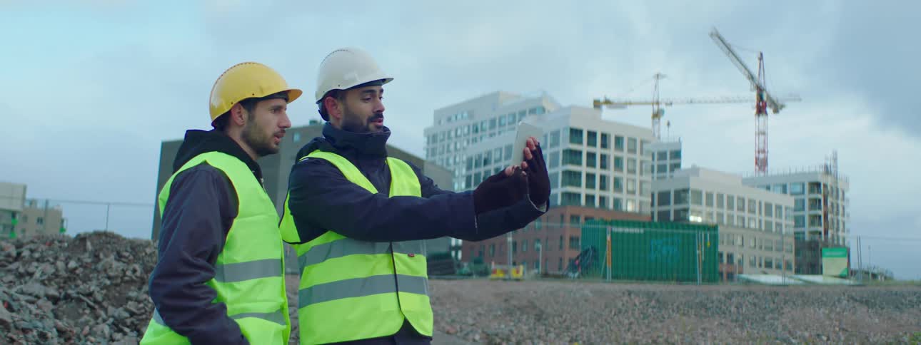 Two construction workers are standing out side near a construction site looking at a cell phone