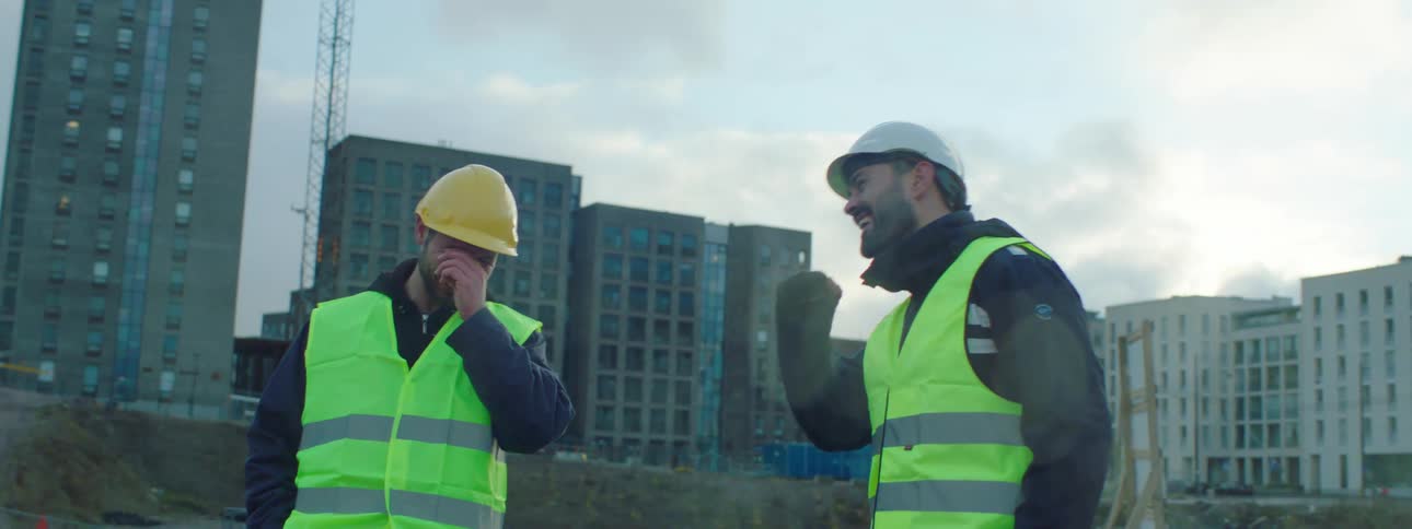 Two male construction workers wearing safety helmets and green vests standing at a construction talking and laughing together
