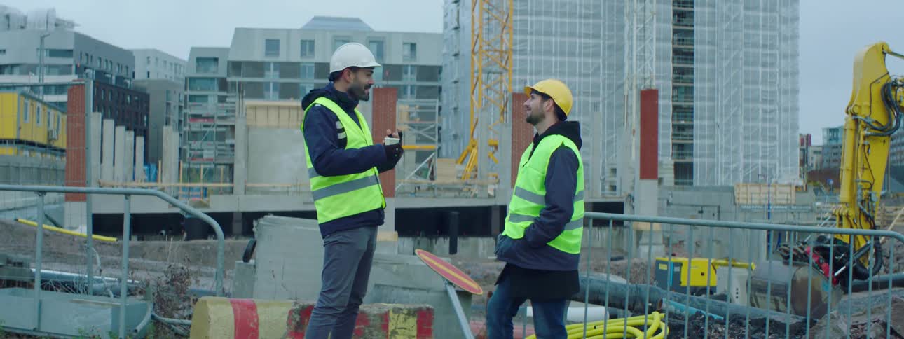 A construction worker holding a cup laughs as he talks with his coworker on a construction site