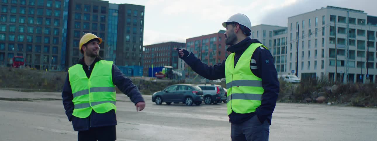 Two male construction workers wearing safety helmets and green vests fist bumping talking and laughing at a parking lot