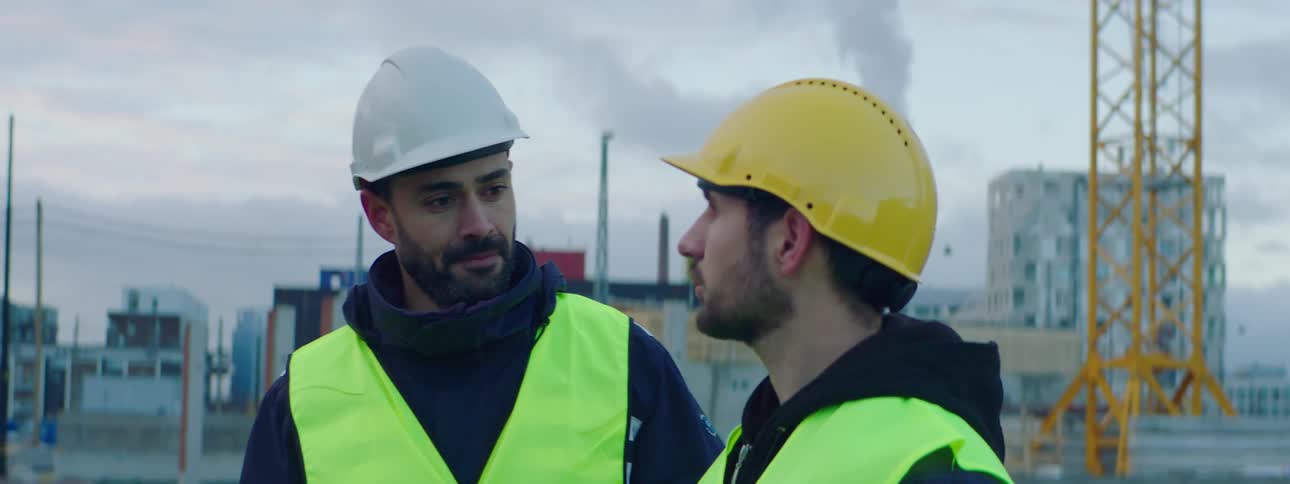 Man wearing a safety helmet and green vest standing at a construction site with a sad looking face while his friends approaches and touches his back in a friendly way