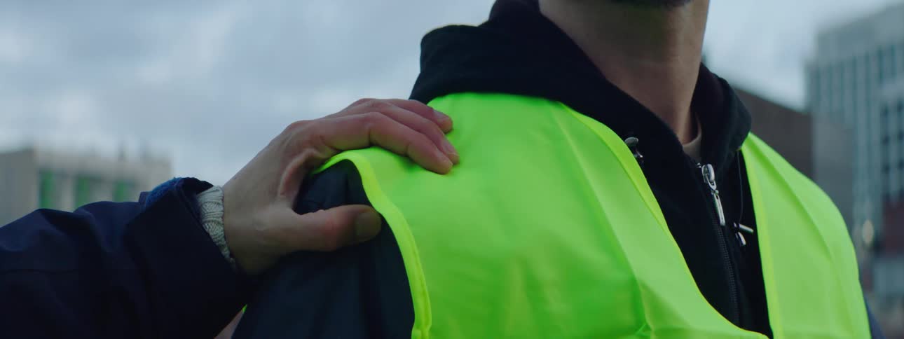 A construction worker stands outside and his coworker puts his hand on his shoulder and he turns and walks