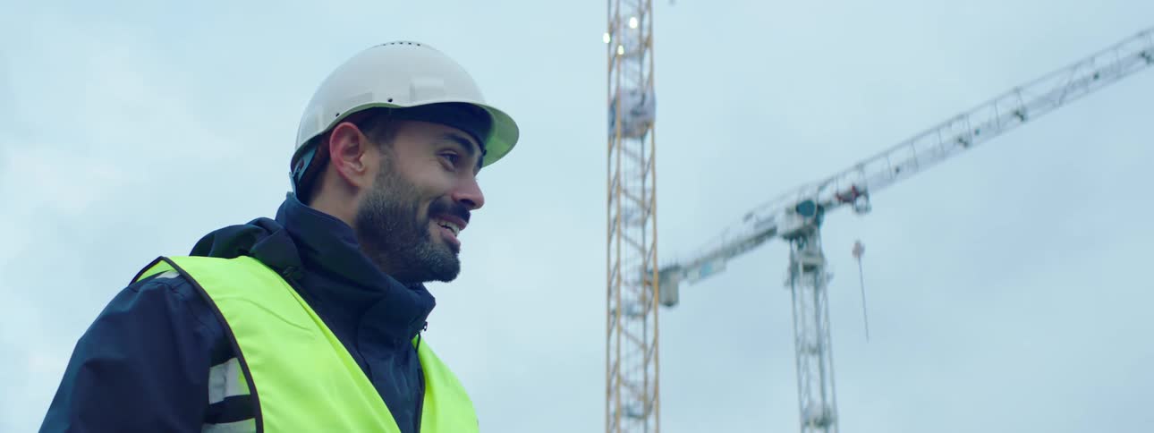 Man wearing a safety helmet coat and a green vest holding a cup while talking and laughing with someone