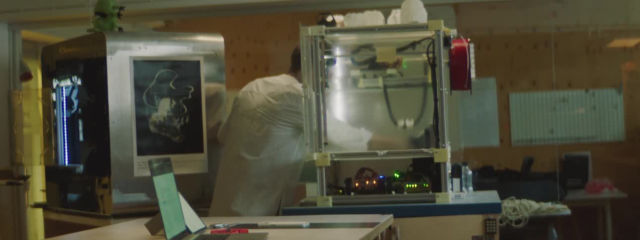 A man wearing a white lab coat opens the doors on a 3d printer and reaches inside