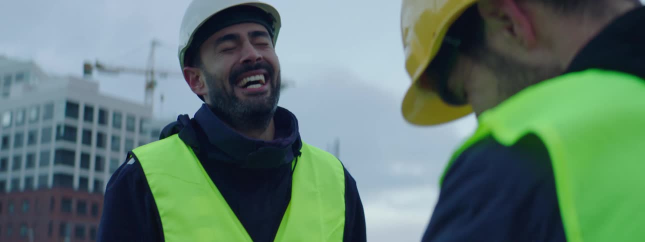 Two construction workers stand in front of a high rise and laugh together