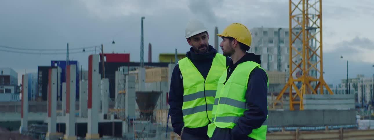 Construction worker wearing safety helmet and vest standing in the middle of the construction site with a sad looking face when his friend approaches and touches his back in a friendly way