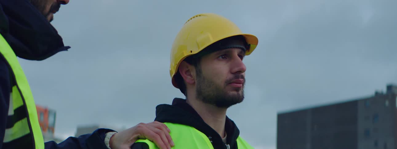 Male construction worker wearing a safety helmet and vest with a introspective face and a man touching his shoulder in a caring way