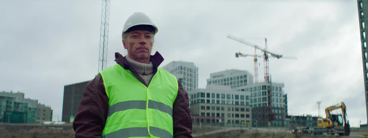 A construction worker is standing outside and there are buildings and cranes behind him