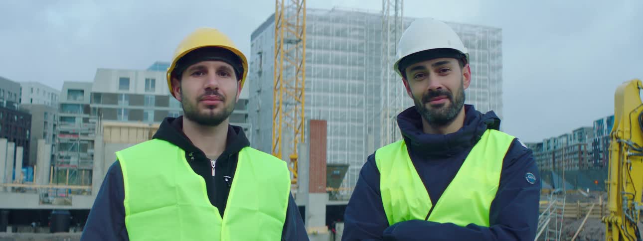 Two male construction workers standing at a construction site side by side wearing safety helmets and green vests
