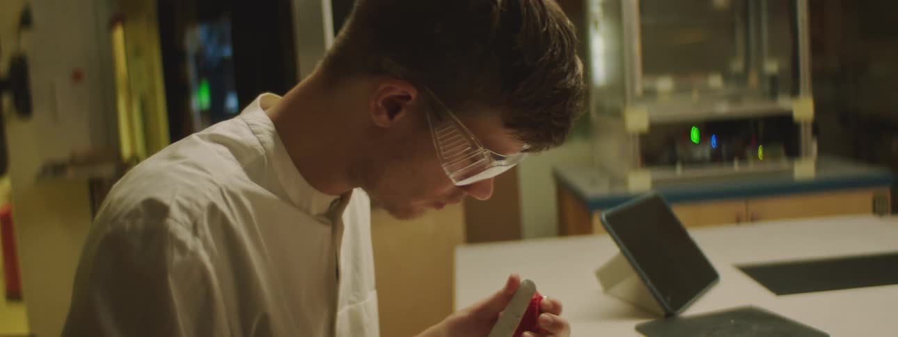 A man wearing safety glasses sitting at a table in a lab using a spatula to shape and smooth a 3d printed model