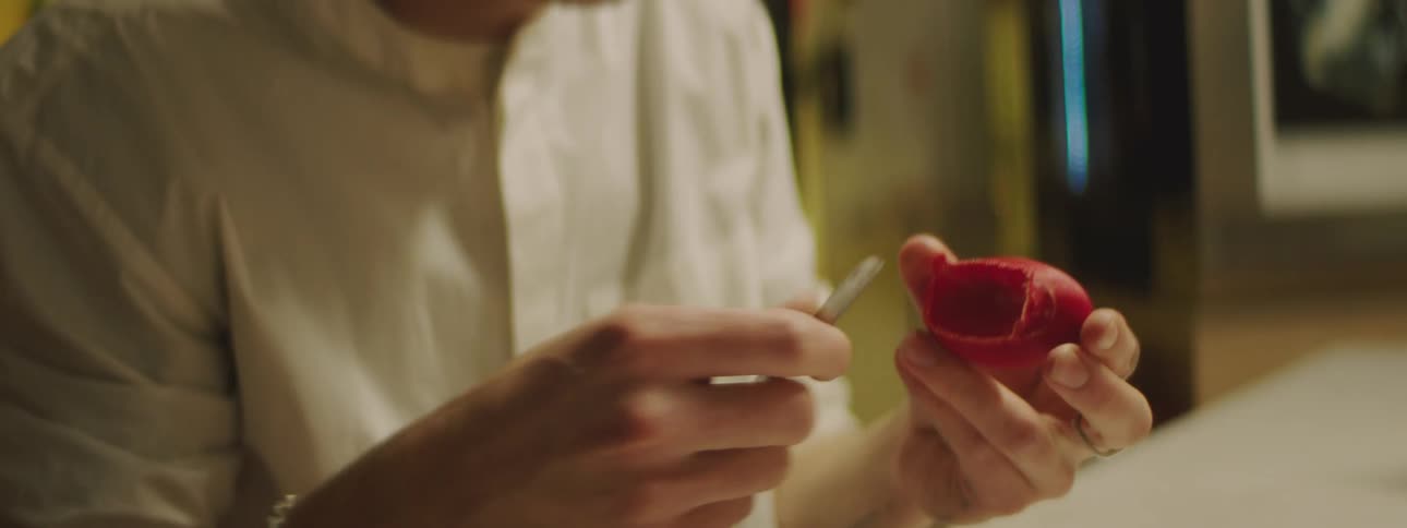 A man is sitting at a table and using a small knife to trim the excess plastic from a red object