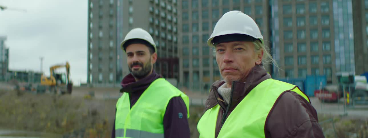 Two construction workers wearing yellow vests and hard hats are standing outside looking ahead