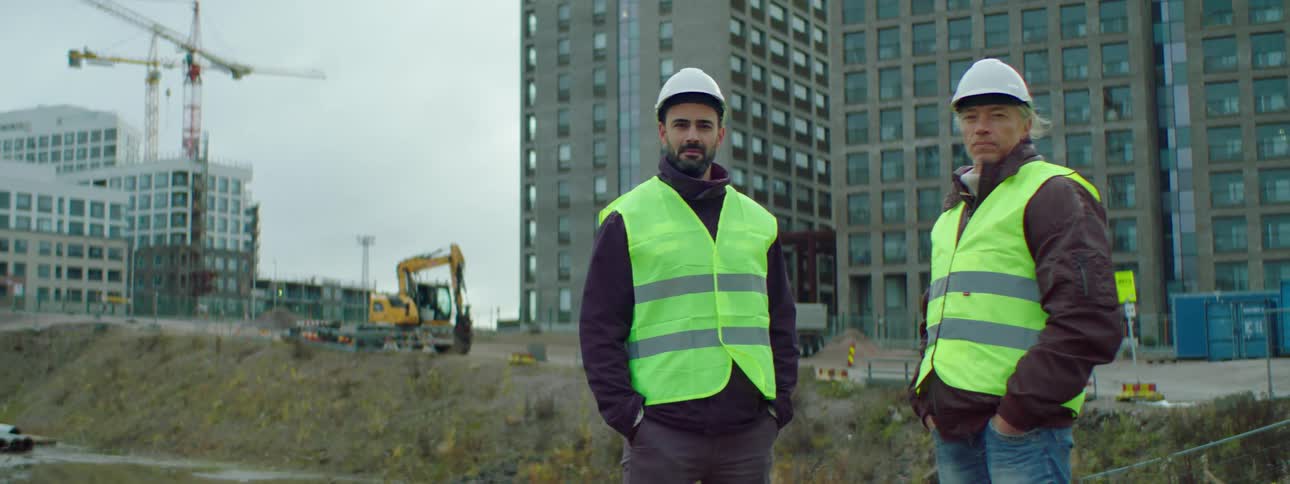 Two construction workers standing with hands in pockets with building and equipment behind them