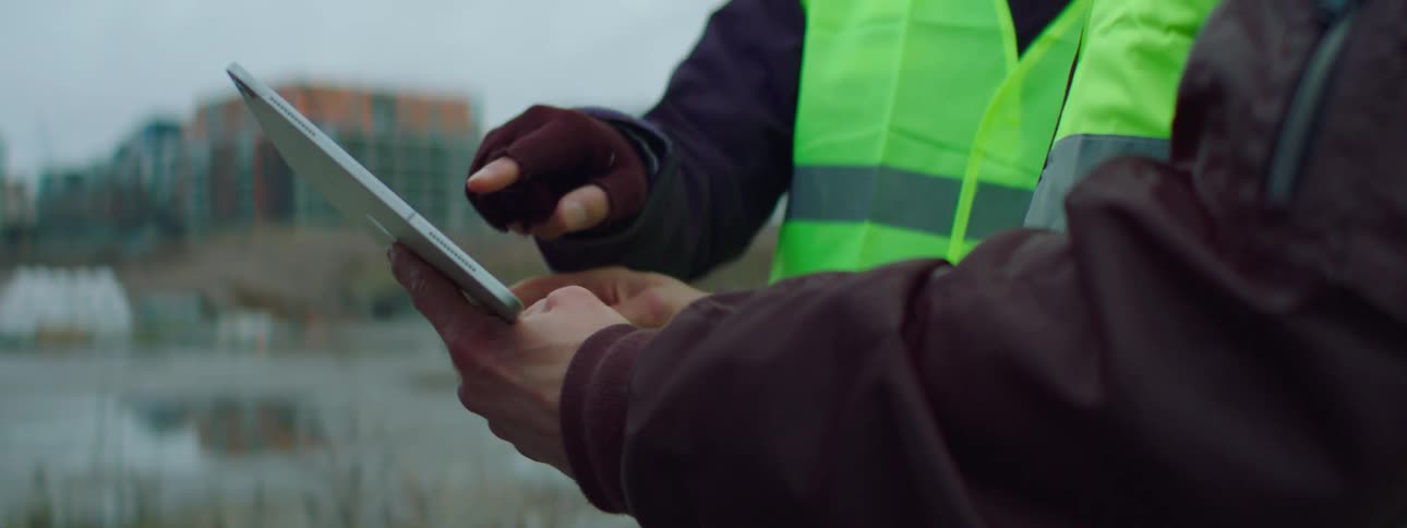 Two male construction workers wearing green vests handling and point at a tablet
