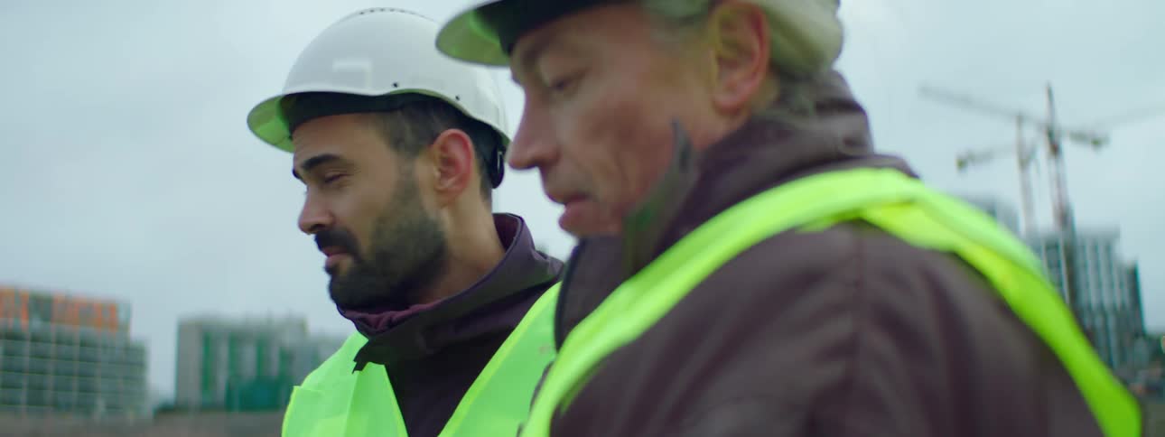 Two male construction workers wearing safety helmets and vests talking at a construction site