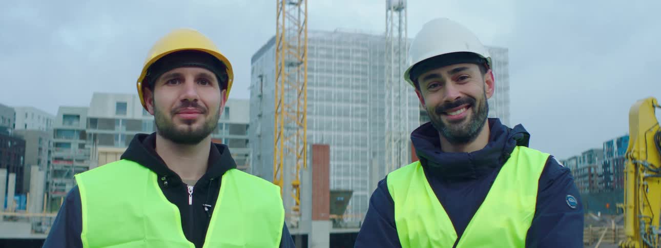 Two construction workers wearing safety helmets and green vests standing at a construction site