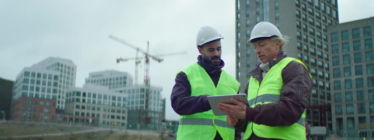 Two construction workers wearing safety helmets and green vests talking and using a tablet in a construction site