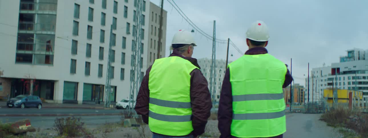Two men wearing safety vests and hats walk next to a construction site in the city