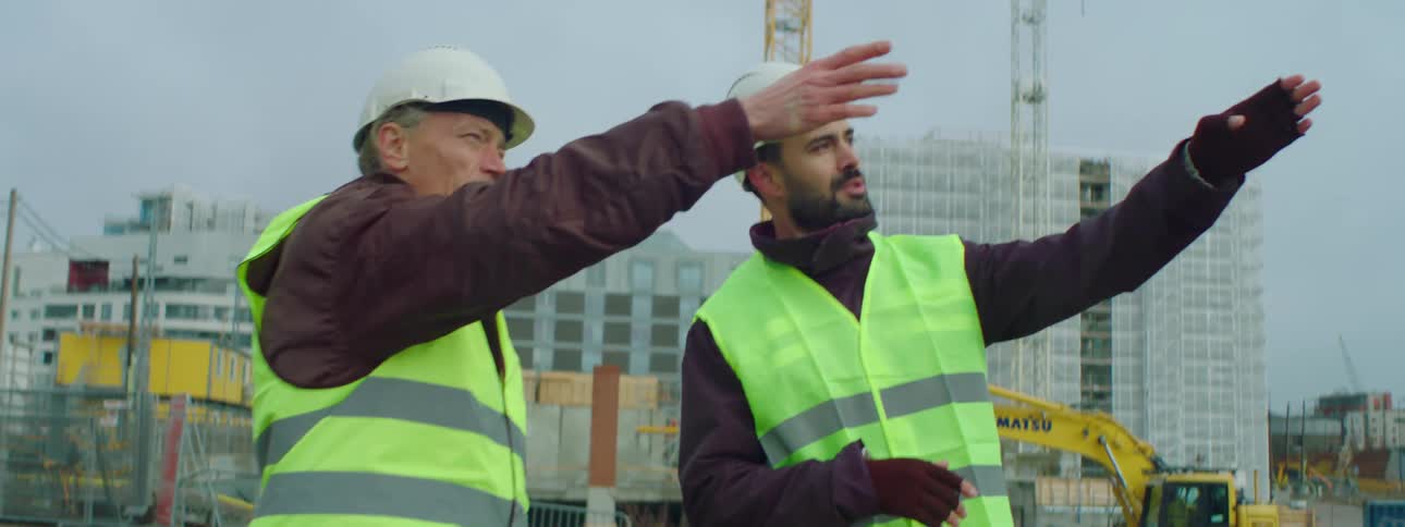 Two male construction workers wearing safety helmets and vests walking together in a construction site talking about the ongoing construction