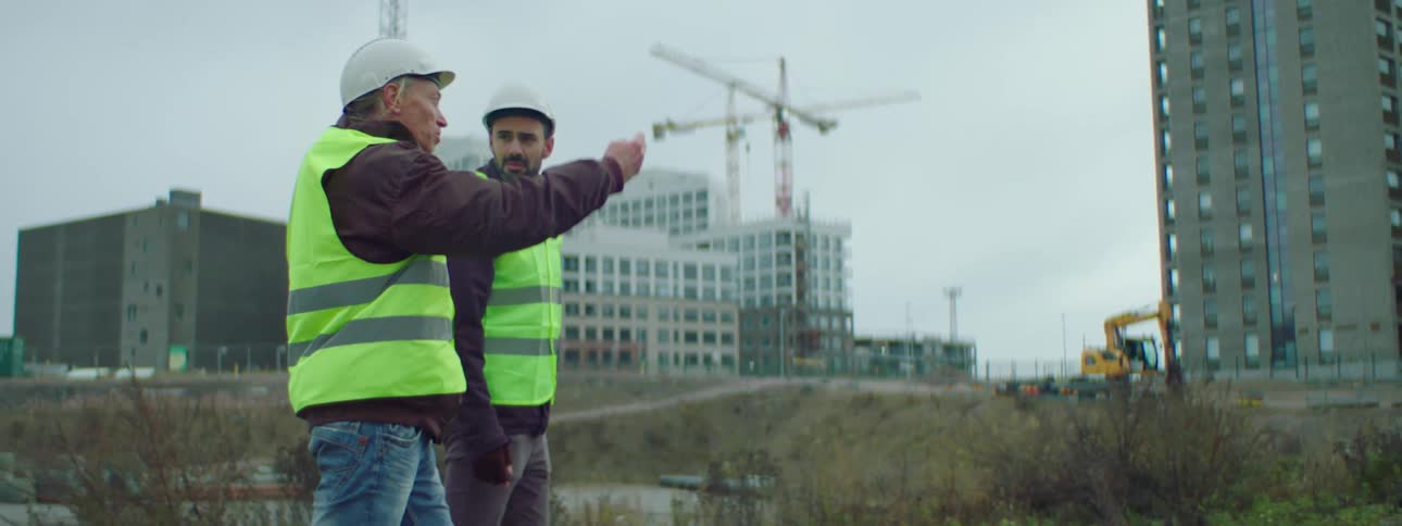 Two male construction workers wearing white safety helmets and vests walking together and talking at a construction site