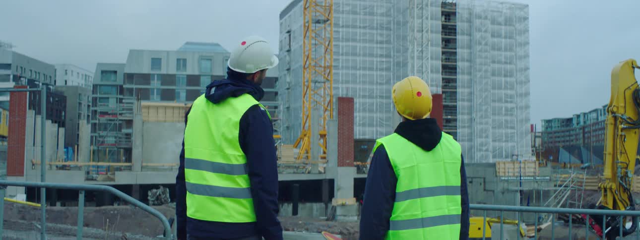 Two male construction workers wearing safety helmets and green vests standing at a construction site pointing and talking about the ongoing construction