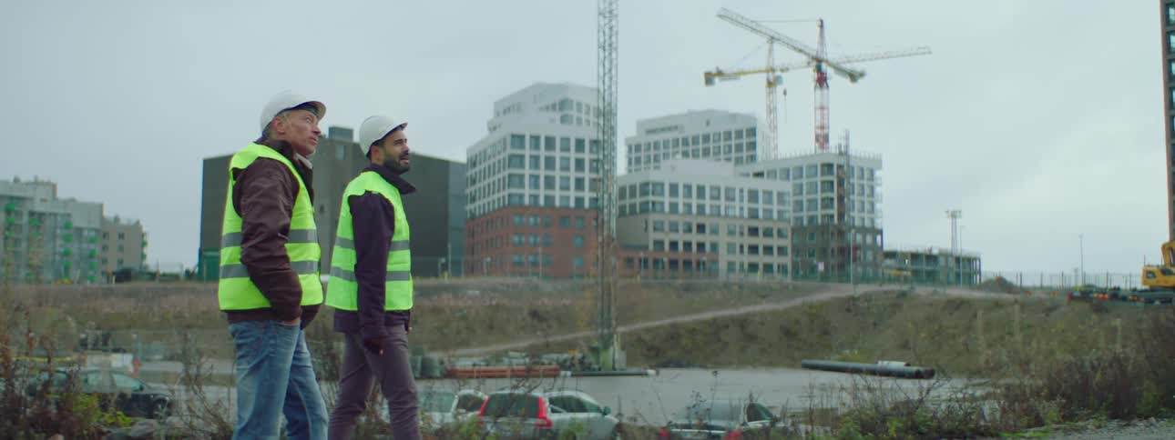 Two constructions workers are walking outside near a construction site and talking as one man points to something