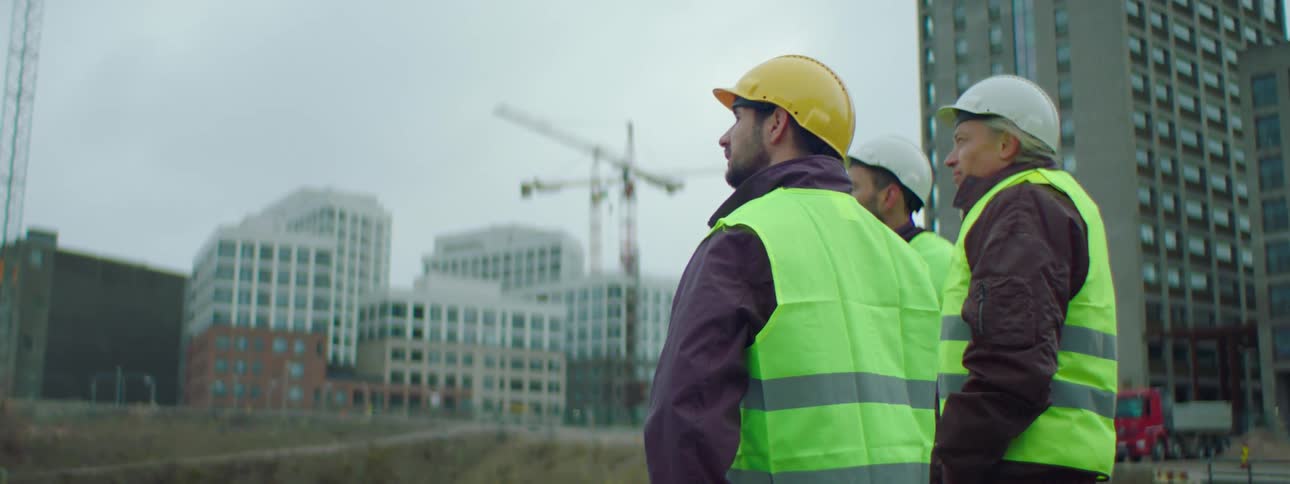 Three construction workers stand talking near a site with a construction crane