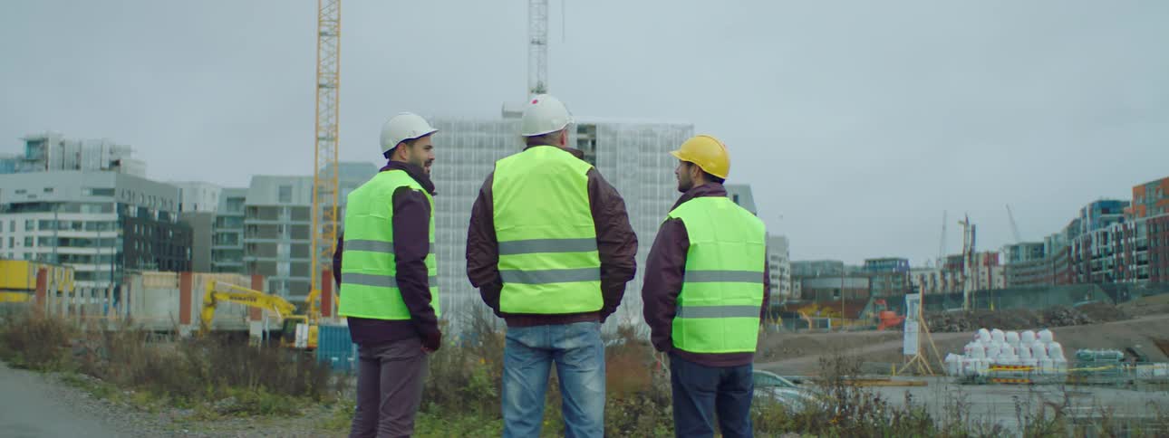 Three construction workers wearing safety helmets and vests standing at a construction site and talking to each other