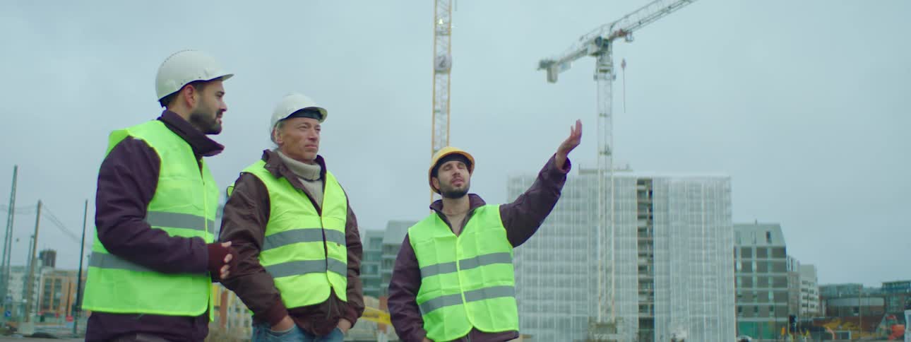 Three male construction workers wearing safety helmets and green vests standing at a construction site talking about the ongoin construction