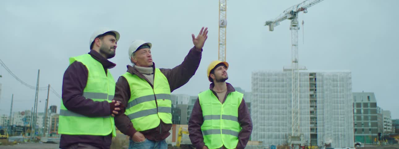 A construction worker points up as he talks to others listening with crane and site behind them