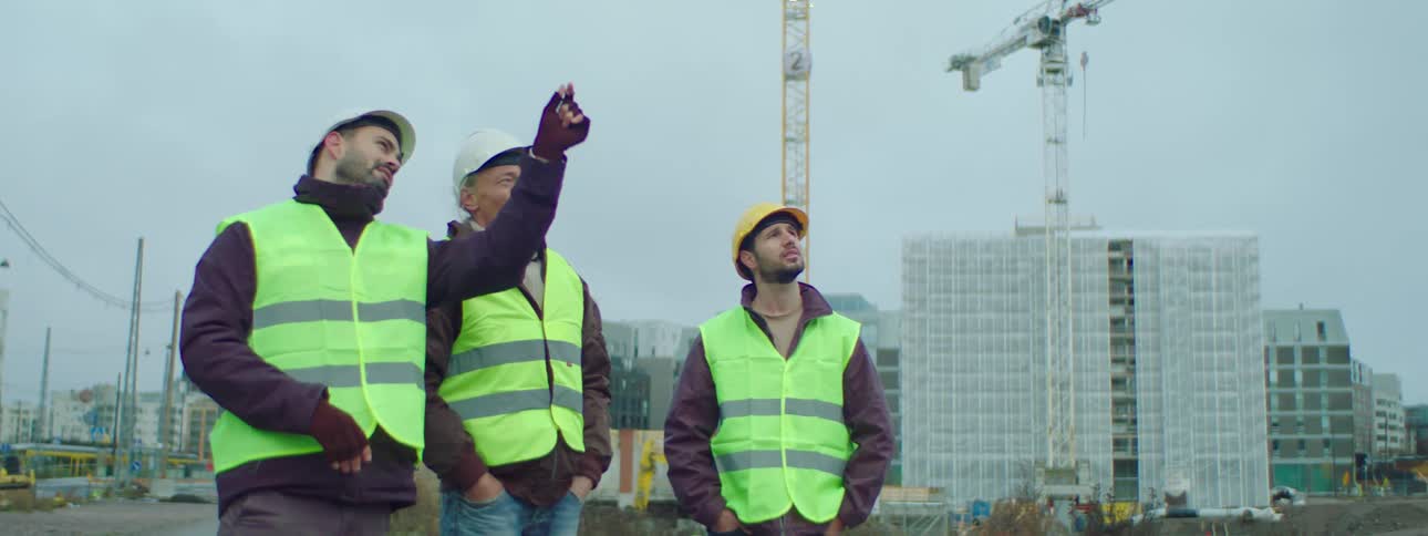 Three male construction workers wearing safety helmets and green vests standing at a construction site talking to each other about the construction