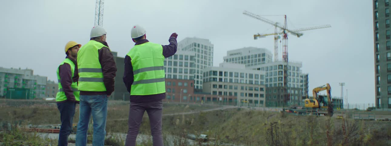 A group of construction workers stand talking while looking at the construction site with cranes and excavator
