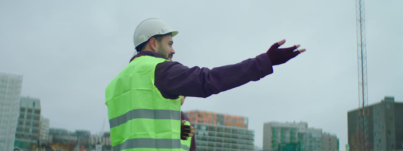 Three male construction workers wearing safety helmets and green vests standing at a construction site and talking about the ongoing construction