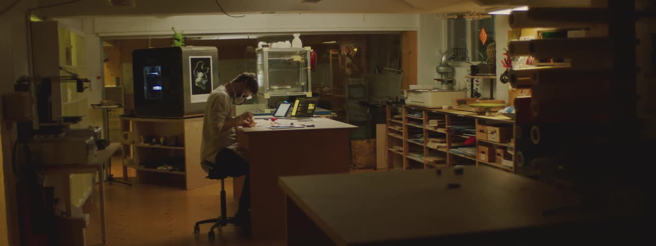 A man is sitting at a desk with a laptop computer and wearing protective goggles and a mask as he mixes something in a small container
