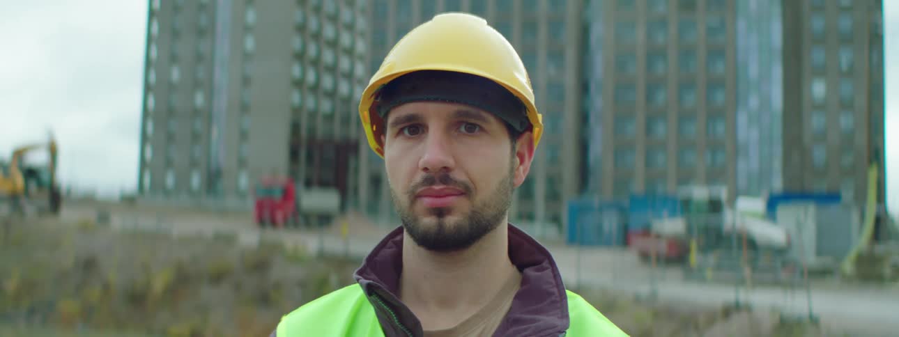 A construction worker stands in front of a building and equipment wearing a hardhat