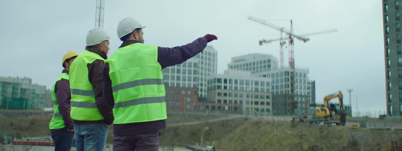Three male construction workers wearing safety helmets and green vests standing at a construction site talking about the ongoing construction