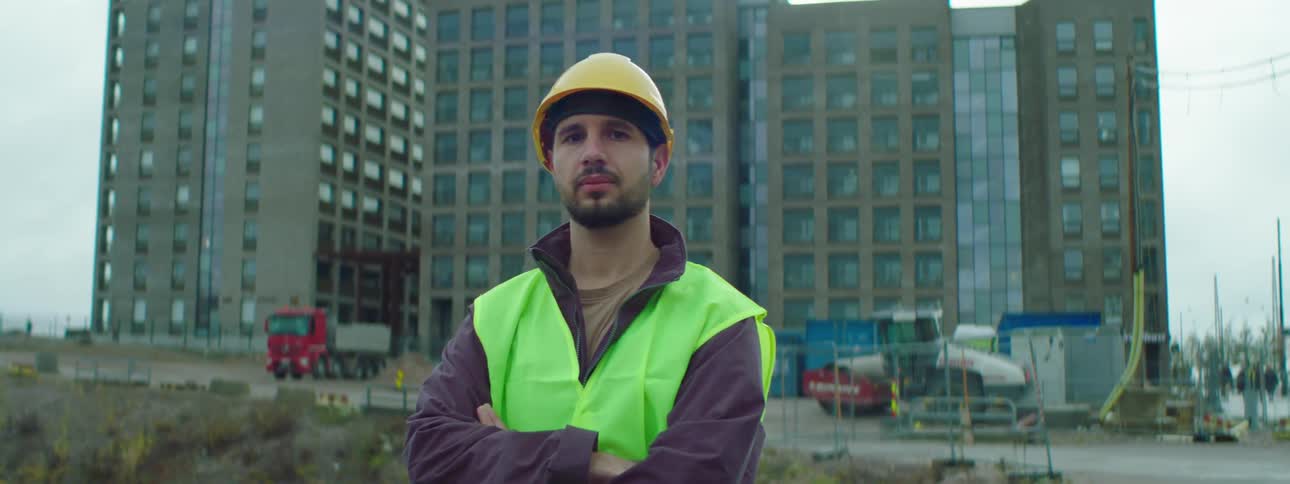 Male construction worker wearing a safety helmet and green vest standing at a construction site with crossed arms