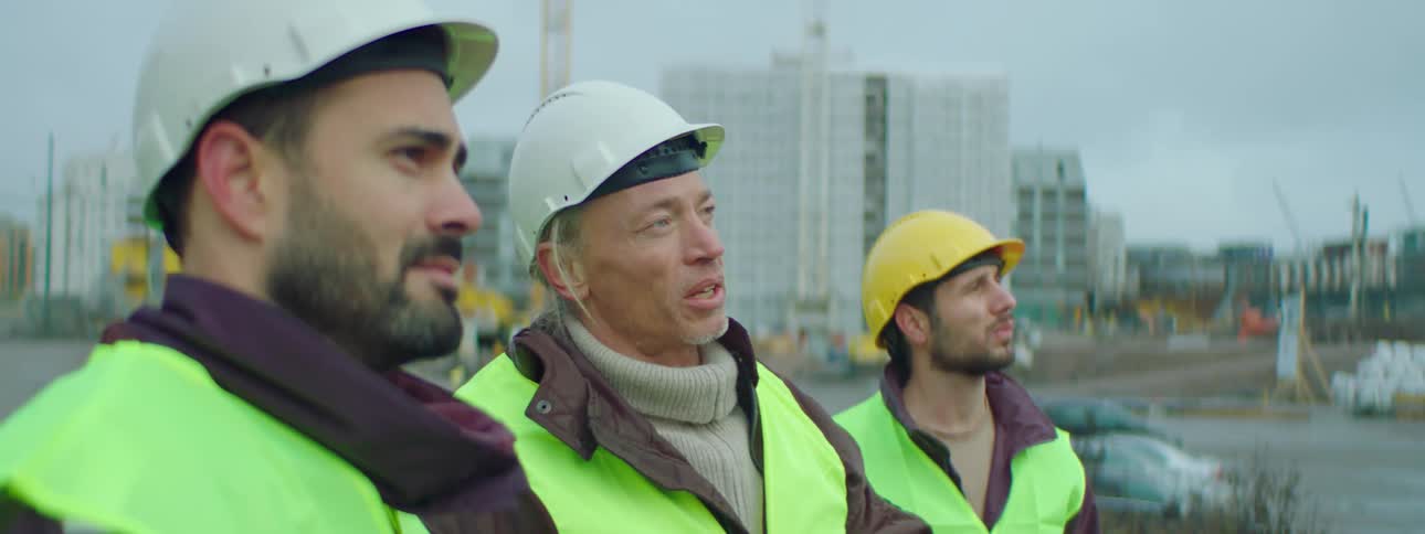 Three construction workers wearing safety helmets and vests talking to each other while standing at a construction site