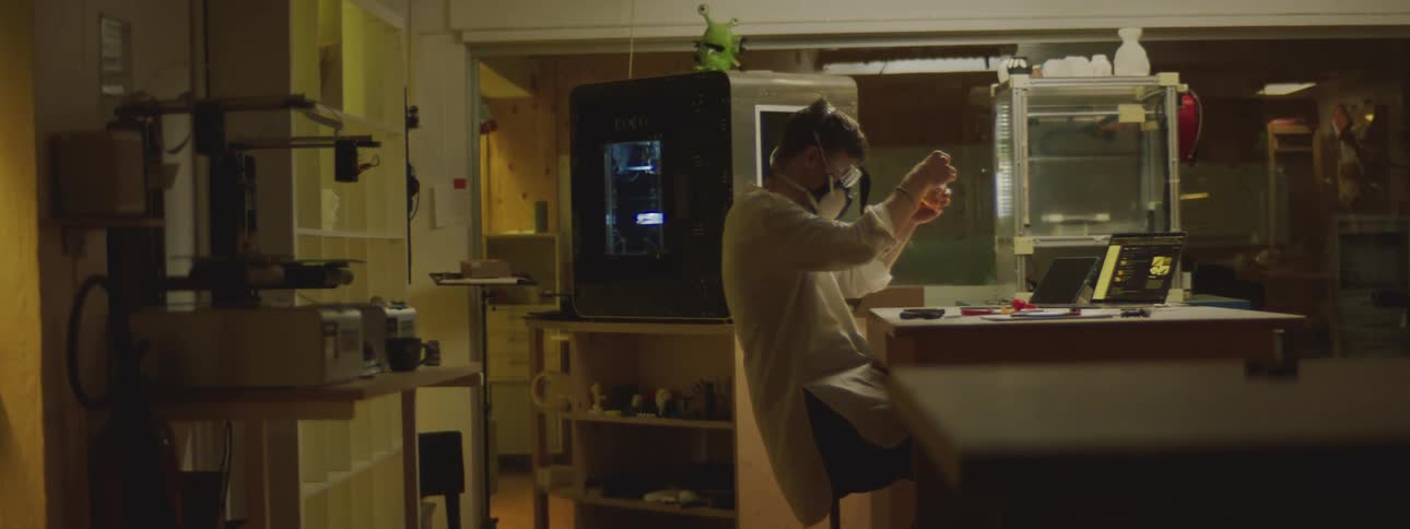 A man is sitting at a desk with a laptop computer and wearing protective goggles and a mask as he mixes something in a small container