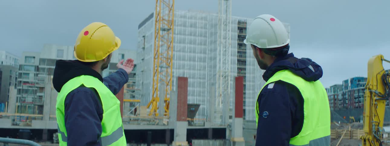 Two construction workers stand in front of a building under construction and point and talk