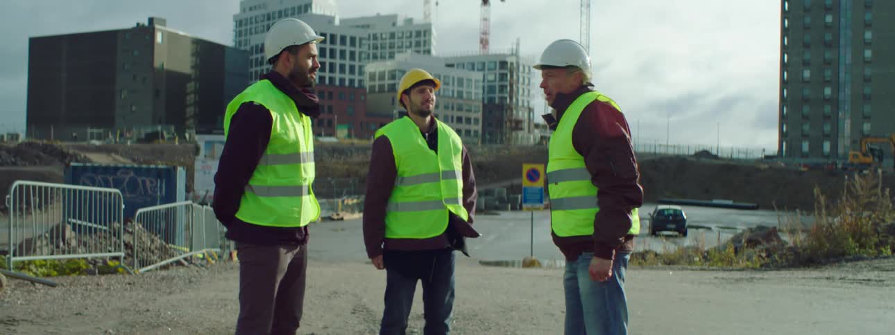 Three construction workers wearing safety helmets jackets and green vests standing in a construction site talking and shaking hands